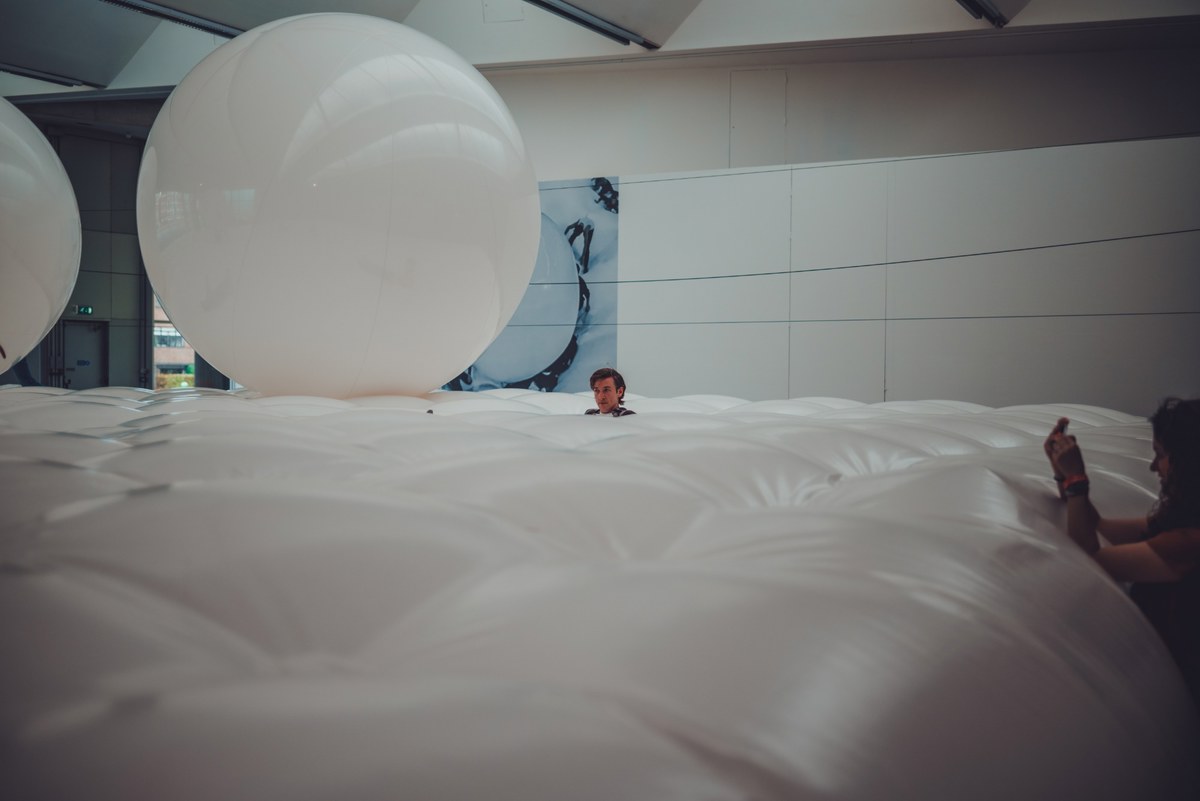 Twee grote witte ballonnen zweven boven een golvend opblaasbaar kunstwerk, waartussen een man opduikt en rechts iemand een foto maakt.