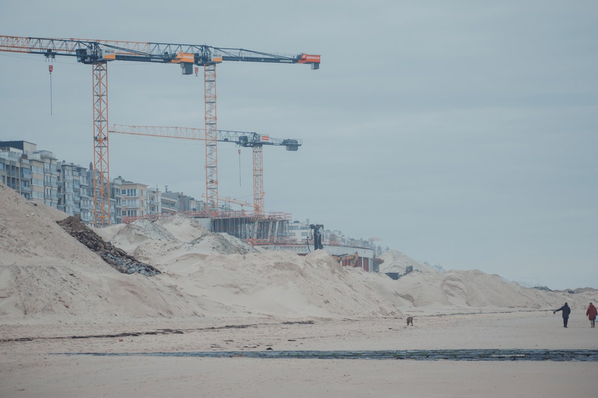 Twee bouwkranen torenen boven appartementsgebouwen en opgeworpen zandbergen uit, terwijl enkele wandelaars over het brede strand lopen onder een grijze lucht.