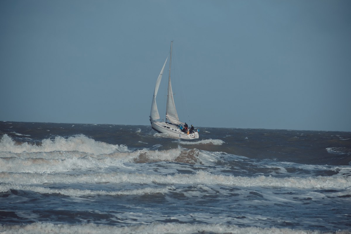 Een zeilboot met bemanning trotseert de ruwe, schuimende golven van de Noordzee onder een bewolkte grijsblauwe hemel bij Middelkerke.