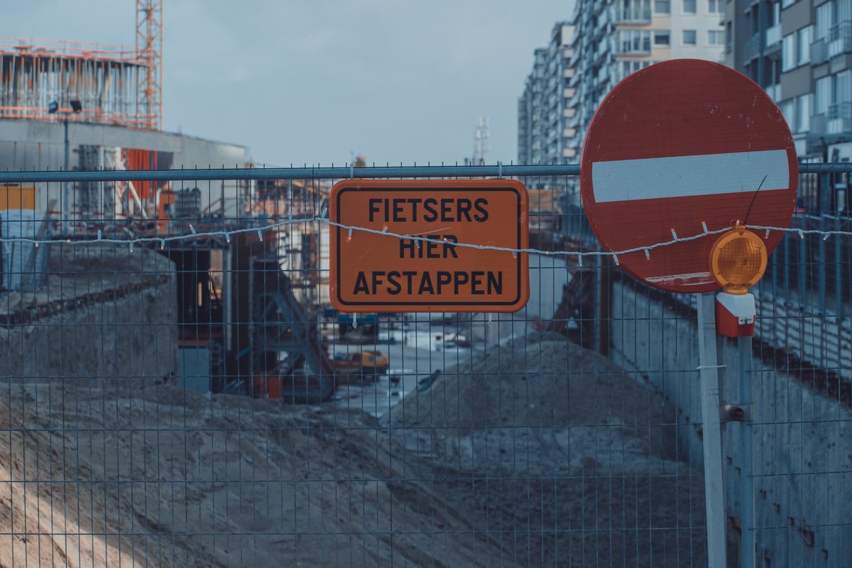 Bij een bouwterrein staan een oranje bord met de tekst Fietsers hier afstappen en een rood verbodsbord langs een hek met hoogbouw op de achtergrond.