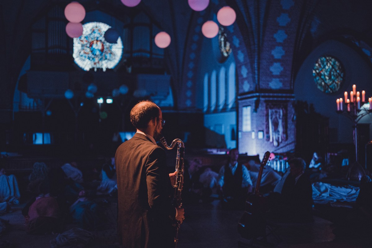 Een saxofonist in pak staat op de rug gezien in een sfeervol verlichte kerk met glas-in-loodramen terwijl bezoekers liggend naar het concert luisteren.