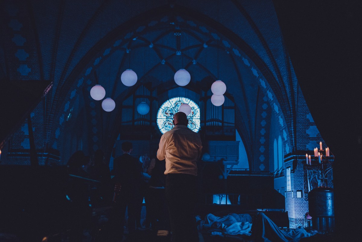 Silhouet van een man in een verduisterde kerk met verlichte bollampen, een gloeiend glas-in-loodraam en kaarsen tijdens een ligconcert.