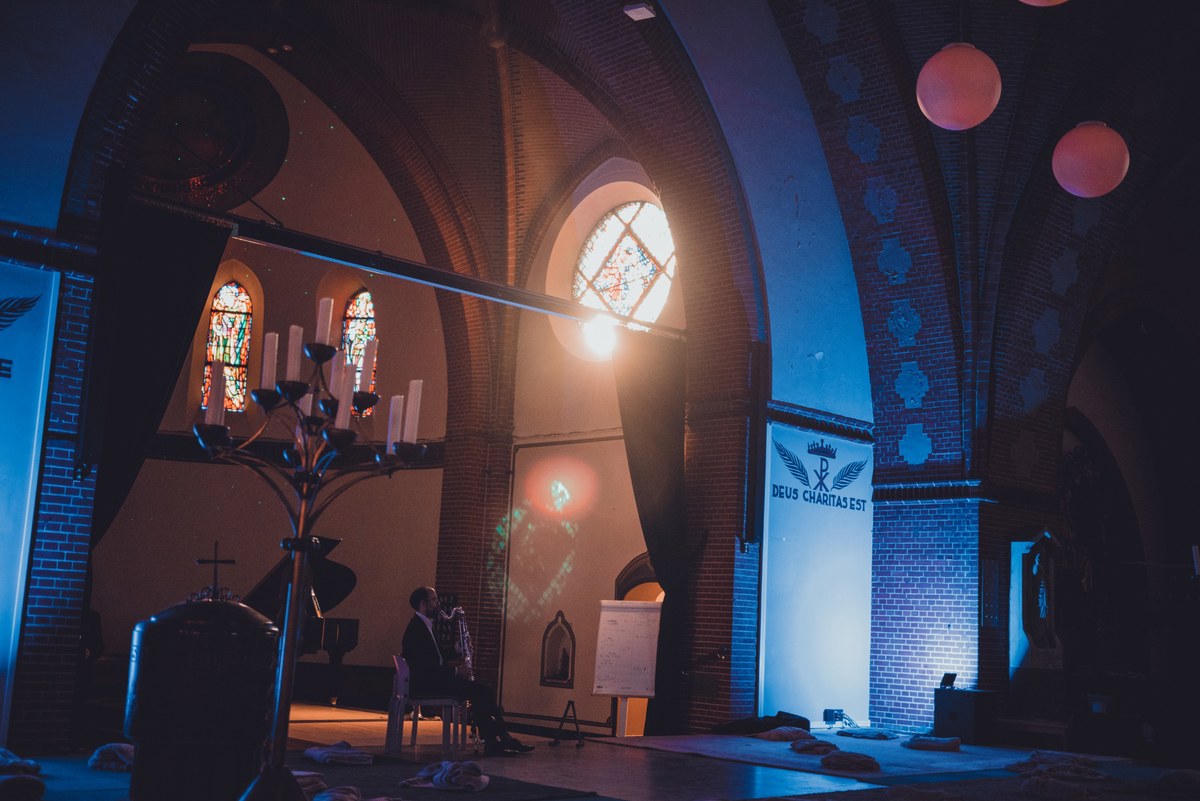 Een pianist speelt in een sfeervol verlichte kerk met blauwe belichting, glas-in-loodramen en een grote kandelaar, terwijl bezoekers op de vloer liggen.