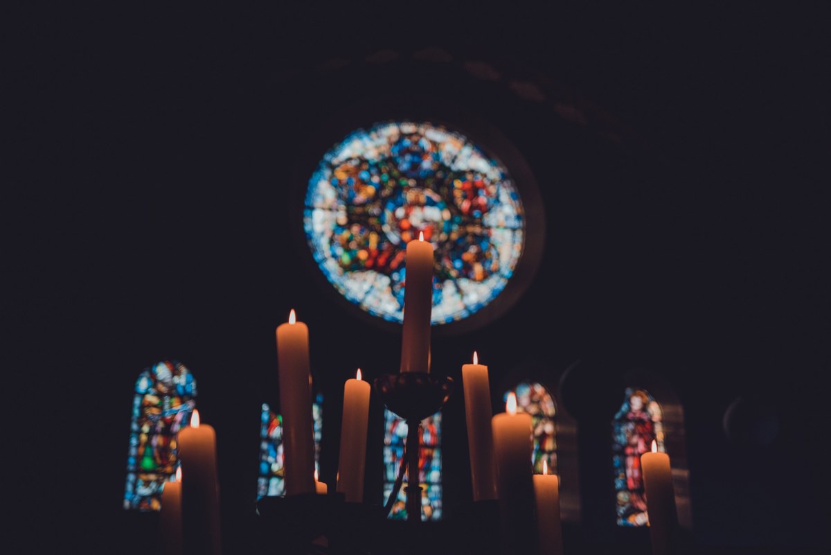 Brandende kaarsen verlichten de voorgrond terwijl een kleurrijk rond glas-in-loodraam oplicht in de donkere ruimte van een kerk.