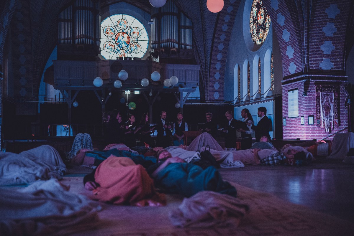 In een sfeervol verlichte kerk met glas-in-loodramen liggen bezoekers onder dekens op de vloer te luisteren naar musici die in het halfdonker optreden.