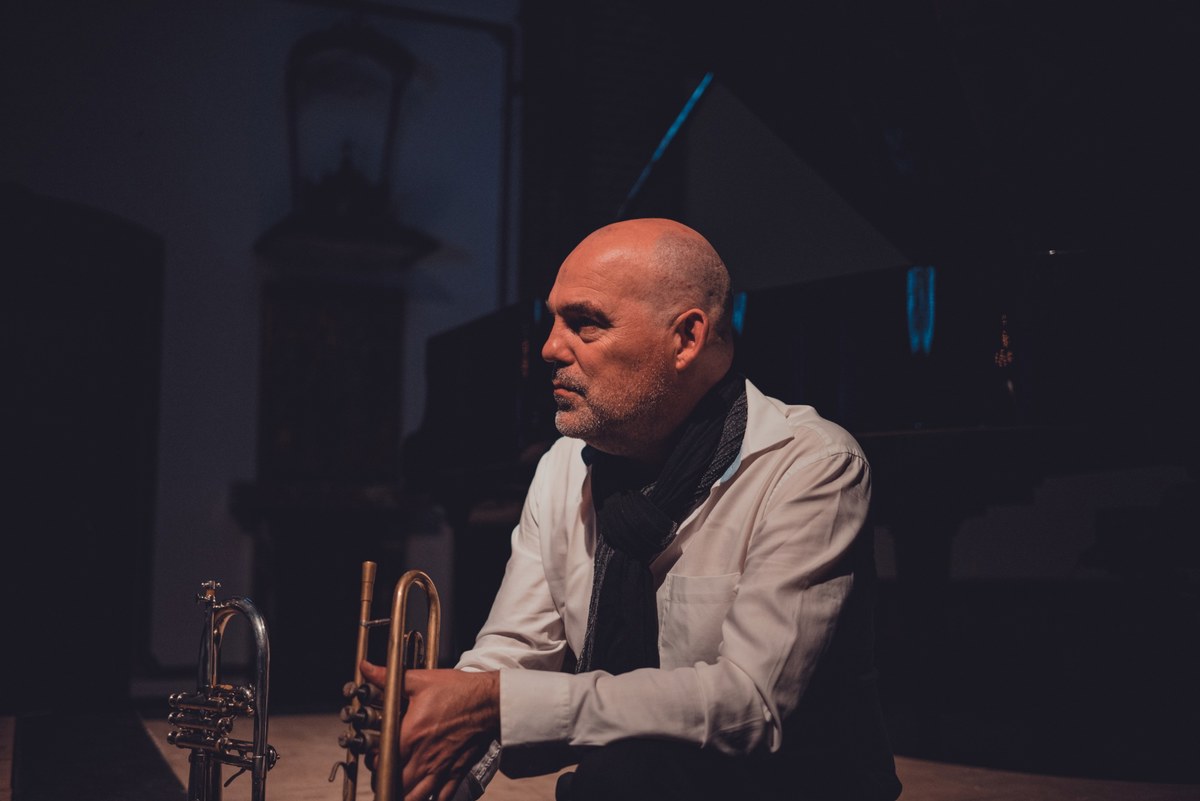 Kale muzikant met sjaal en wit overhemd zit in gedempt licht met een trompet in zijn handen voor een piano.