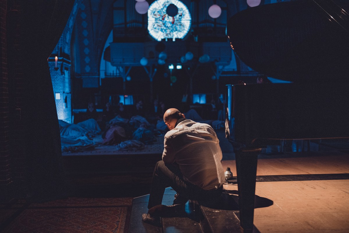 Een pianist zit gebogen achter een vleugel in een donkere zaal, terwijl bezoekers op de vloer liggend luisteren onder blauwe projecties.