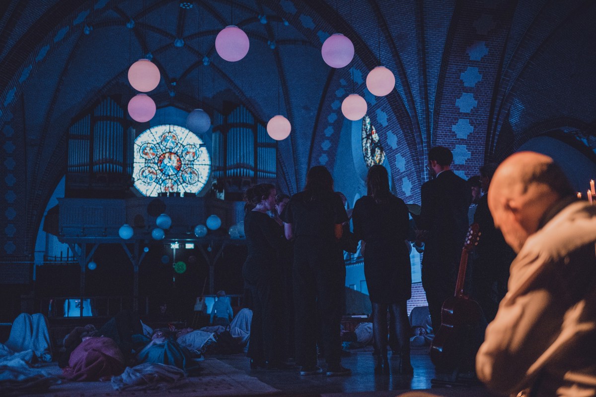 Bezoekers staan te praten in een sfeervol verlichte kerk met glas-in-loodraam, orgel en verlichte ballonnen, terwijl mensen op de grond liggen te luisteren.