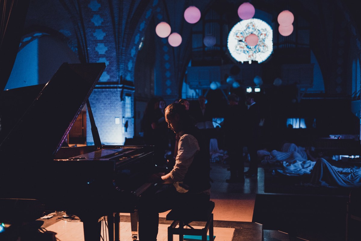Pianist speelt op een vleugel in een sfeervol verlicht kerkgebouw terwijl bezoekers liggend op de grond in het schemerdonker meeluisteren.