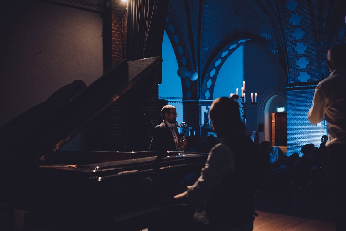 Saxofonist speelt in een sfeervol verlichte kerk met blauwe gewelven, gezien vanachter een vleugel en silhouetten van luisterende bezoekers.