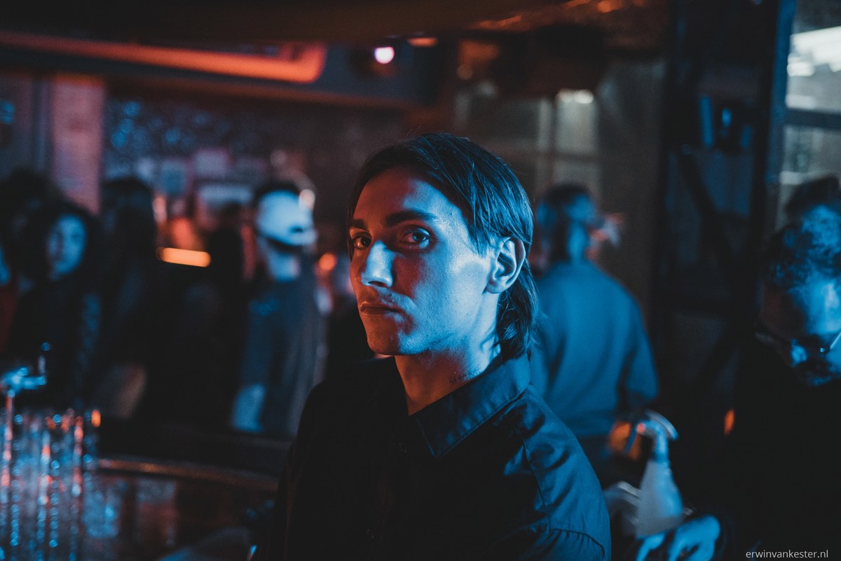 Jonge man met halflang donker haar en zwart overhemd kijkt opzij in een donkere club, verlicht door blauw en rood licht met mensen op de achtergrond.