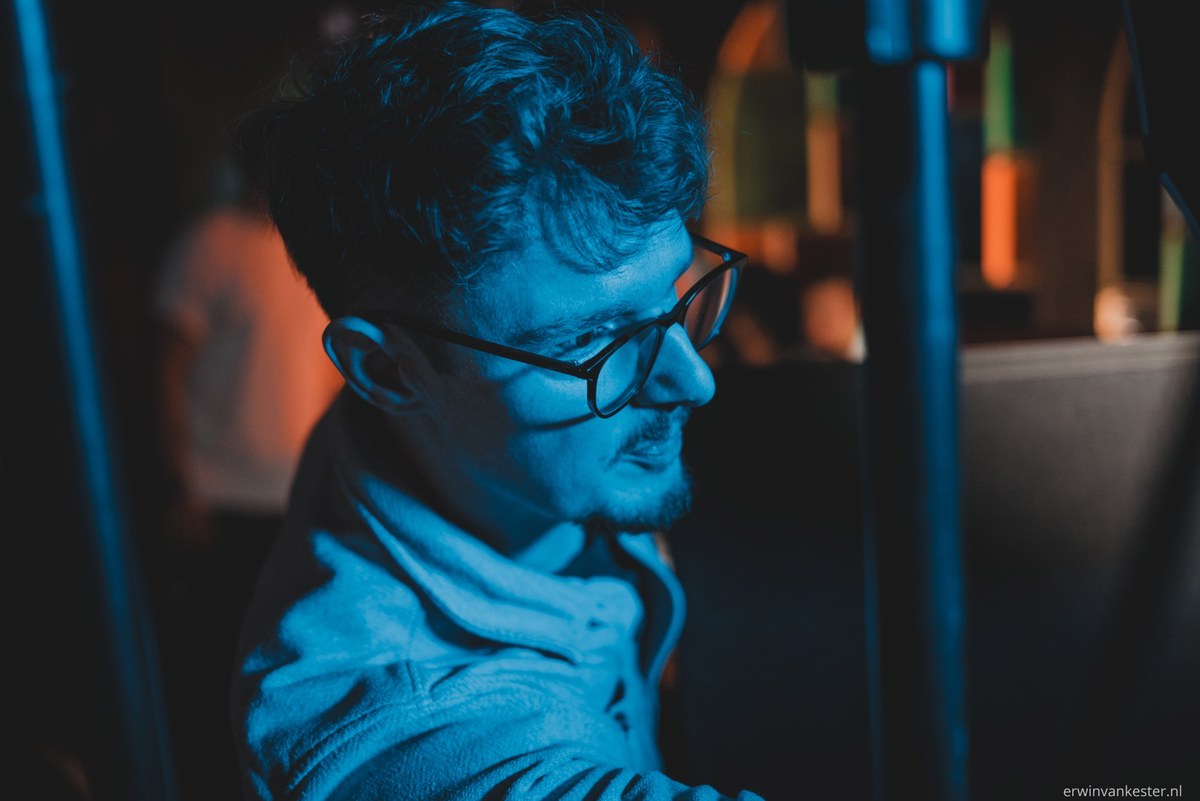 Man met bril en krullend haar in profiel, badend in blauw licht met warme oranje accenten op de achtergrond tijdens een commercial-opname.