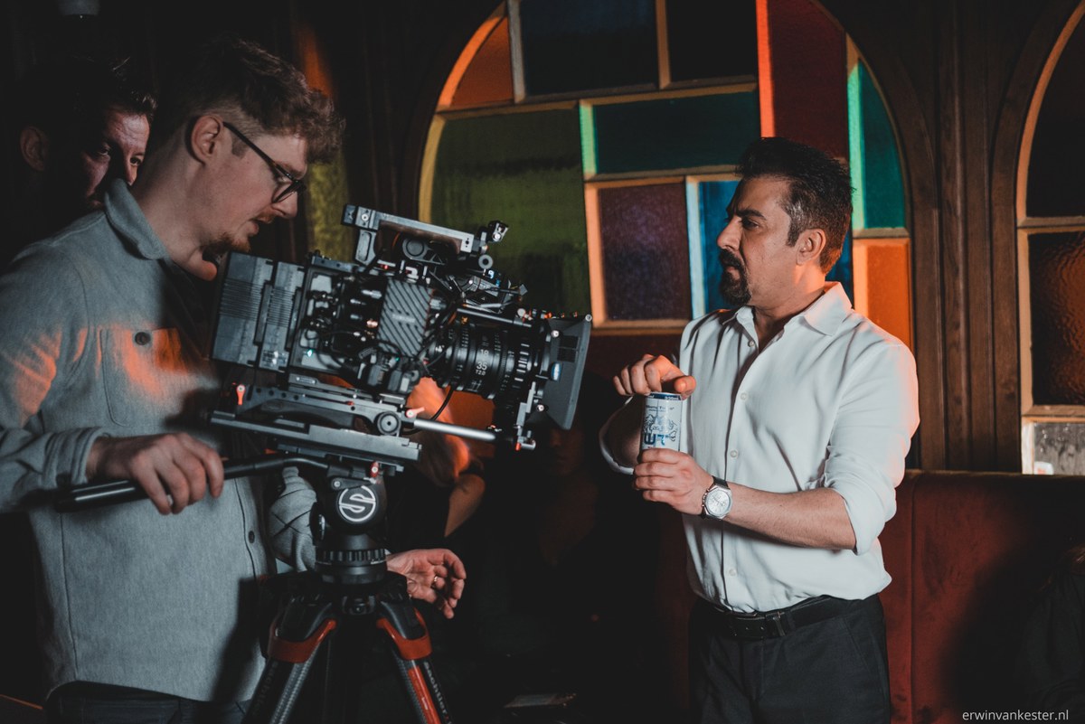 Cameraman bedient een professionele filmcamera op statief terwijl een pratende man met blikje ernaast staat, voor een kleurrijk glas-in-loodraam.