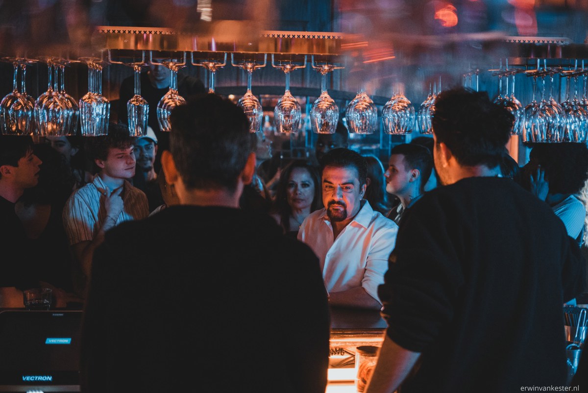 Achter een bar met rijen omgekeerde wijnglazen praten mensen in warm en blauw licht, terwijl twee mannen op de voorgrond in silhouet staan.