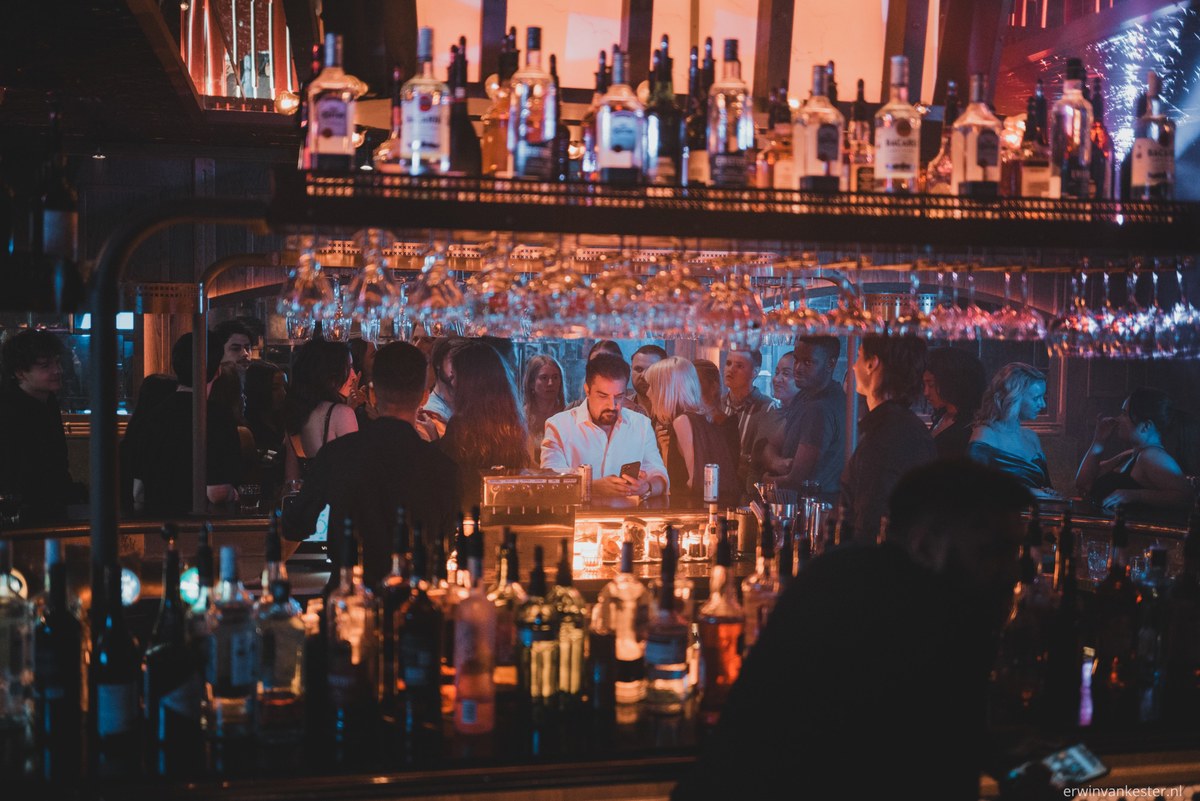 Barman kijkt op zijn telefoon achter een goedgevulde bar terwijl gasten in het warme, sfeervolle licht van een drukke cocktailbar staan.