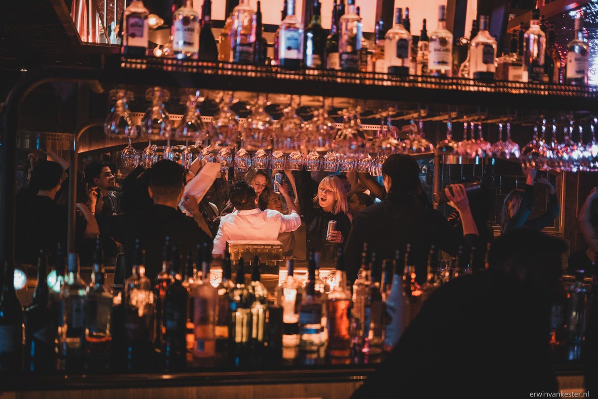 Bezoekers kletsen en drinken aan een sfeervolle bar met warme verlichting, omringd door talloze flessen en boven hen hangende wijnglazen.