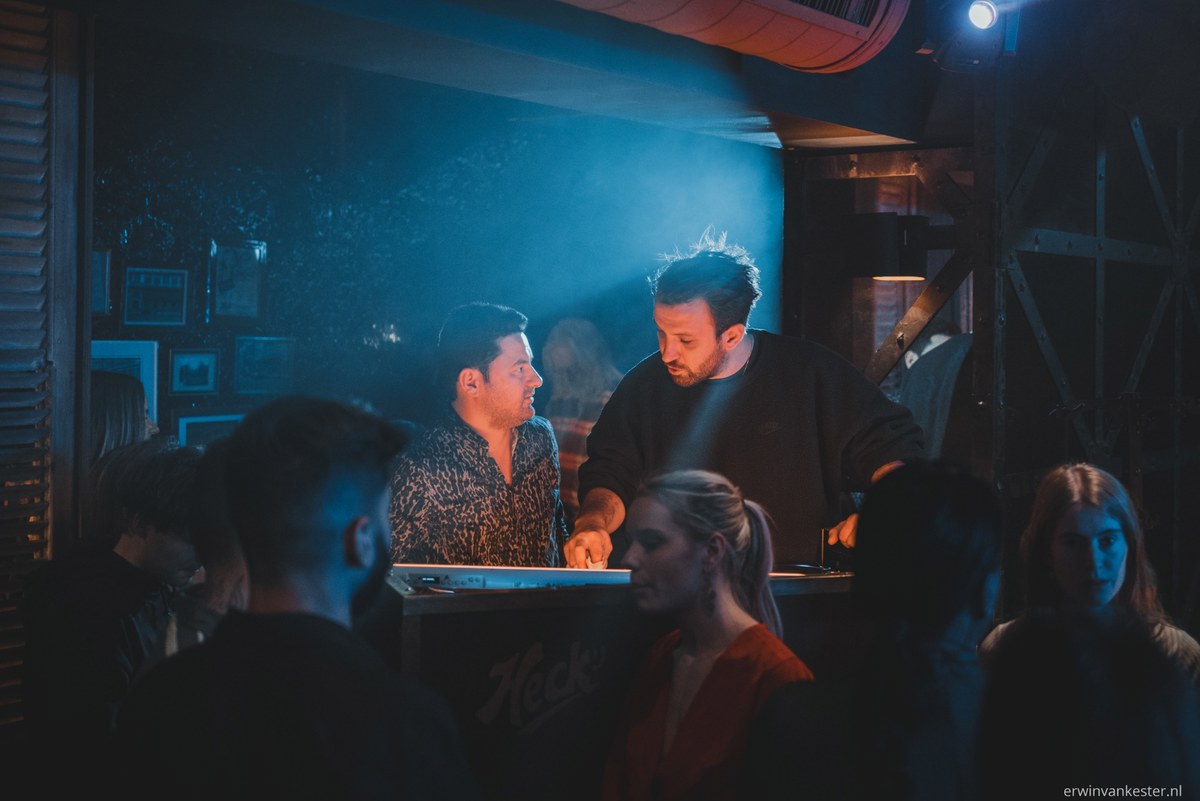 Twee mannen achter een DJ-booth in een sfeervolle, blauw verlichte club terwijl bezoekers op de voorgrond staan te dansen.