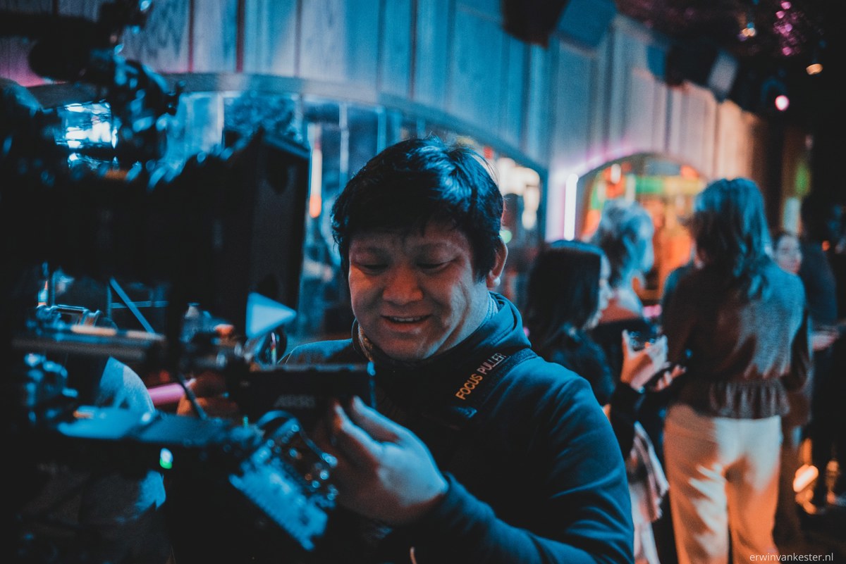 Cameraman bedient een professionele filmcamera tijdens een opname in blauw en roze aangelicht decor, terwijl op de achtergrond crewleden aan het werk zijn.