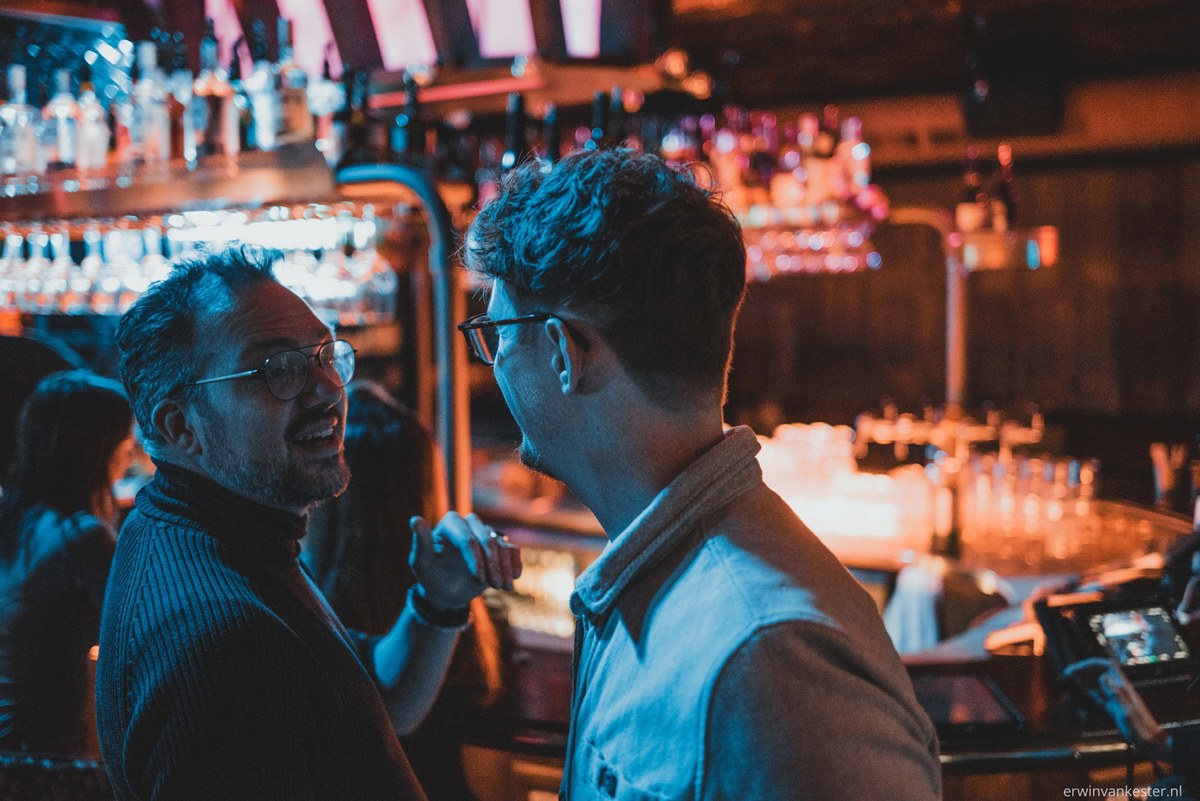 Twee mannen met bril praten geanimeerd met elkaar aan een bar die baadt in blauw en oranje neonlicht, met flessen op de achtergrond.