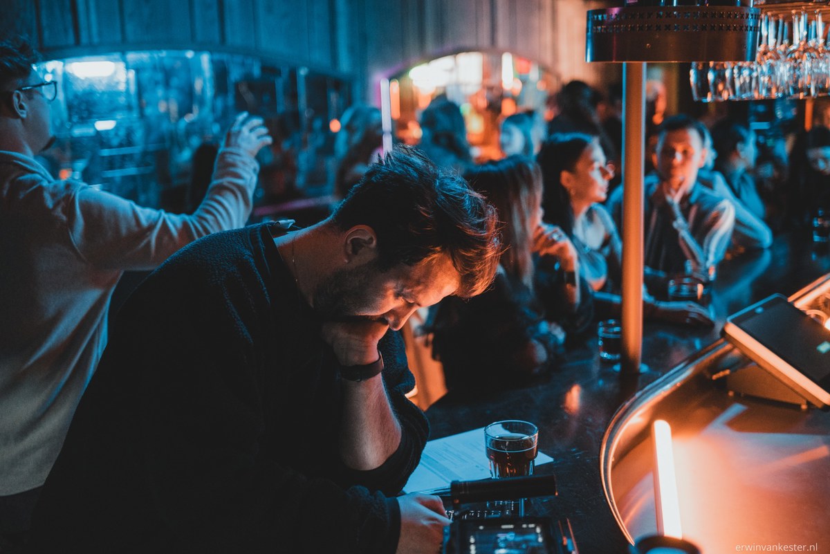 Man leunt voorovergebogen aan een bar met een glas bier, terwijl op de achtergrond mensen in blauw en oranje licht samenkomen.