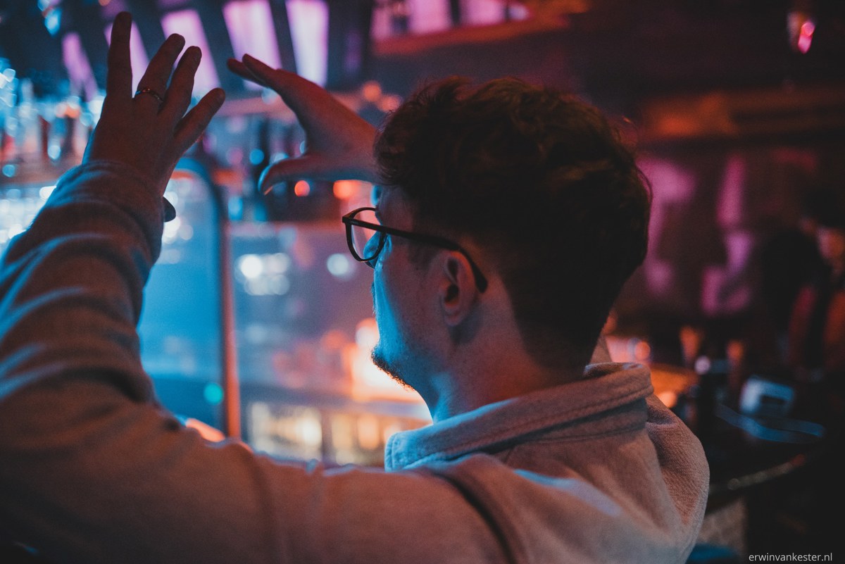 Man met bril en baard van achteren gezien, met opgeheven handen, tegen een sfeervolle achtergrond met blauwe en roze verlichting.