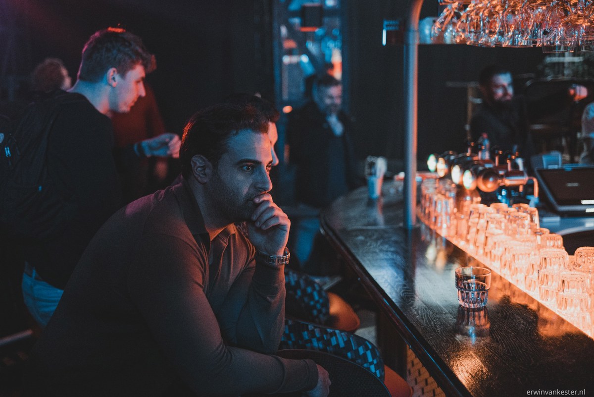 Man met baard zit peinzend aan een sfeervol verlichte bar terwijl op de achtergrond meerdere mensen in blauw-oranje licht met elkaar praten.