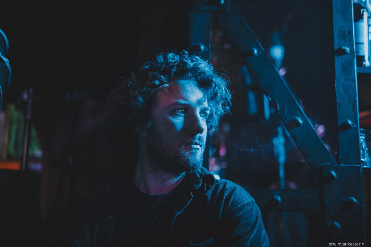 Man met krullend haar en baard kijkt zijdelings weg, badend in blauw en paars licht tussen industriële stalen constructies op een filmset.
