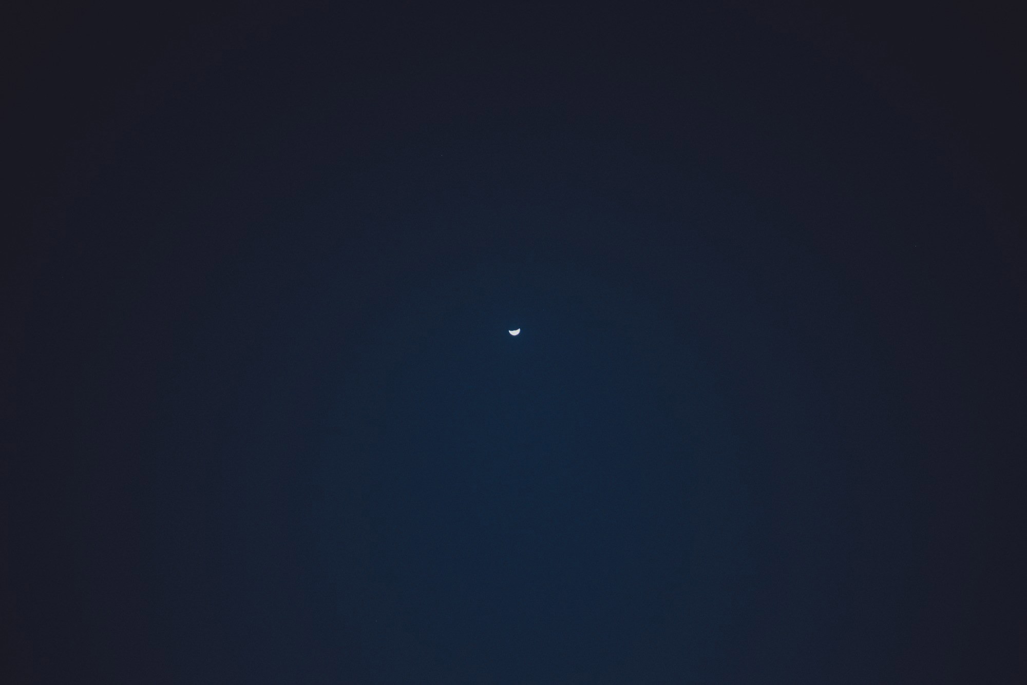 Een dunne sikkelmaan hangt als klein lichtpuntje midden aan een diepblauwe, bijna zwarte nachthemel boven Funchal.