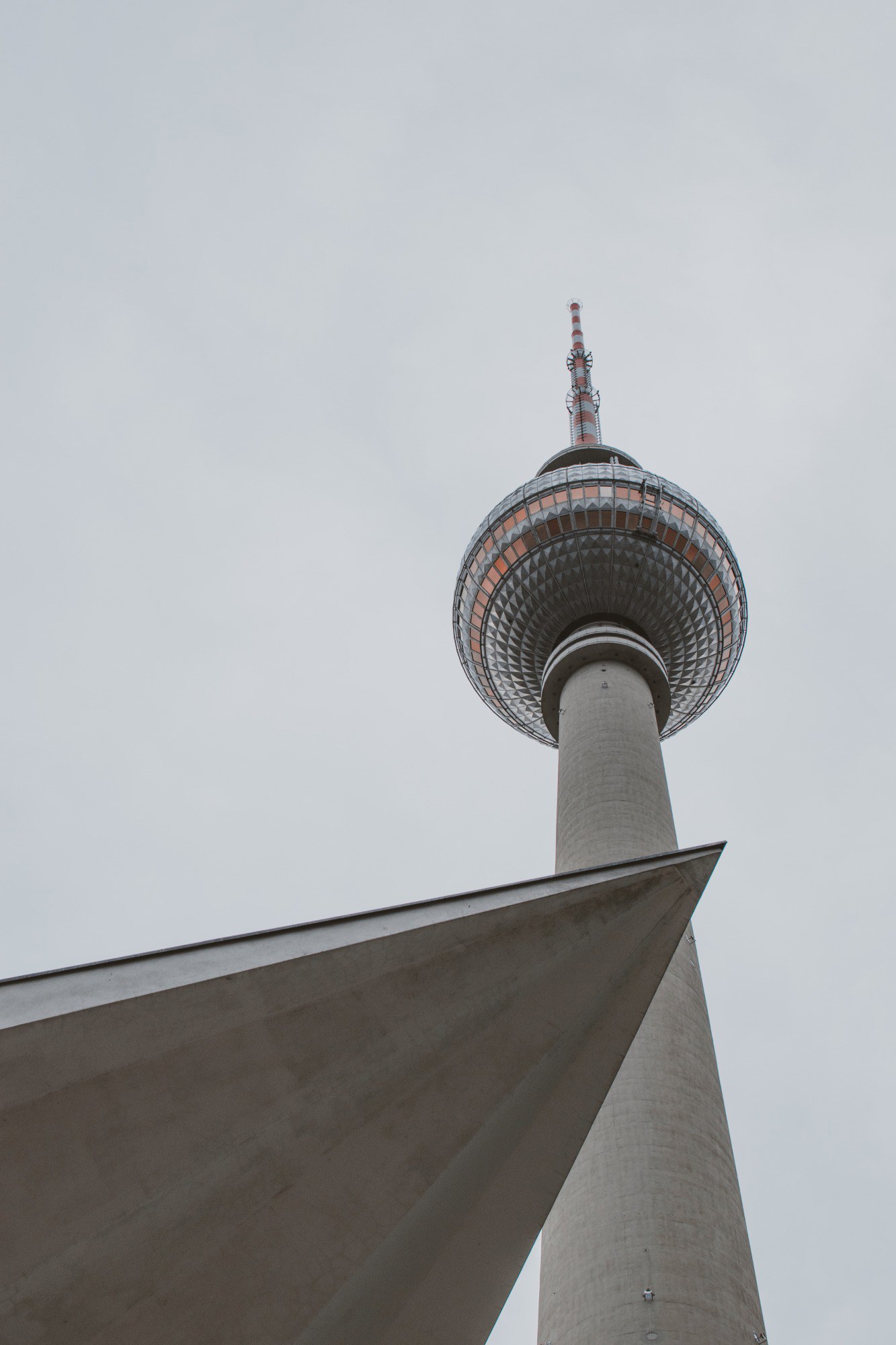 De Berlijnse Fernsehturm met zijn karakteristieke bol torent hoog boven een scherp geometrisch betonnen dak uit tegen een grijze lucht.
