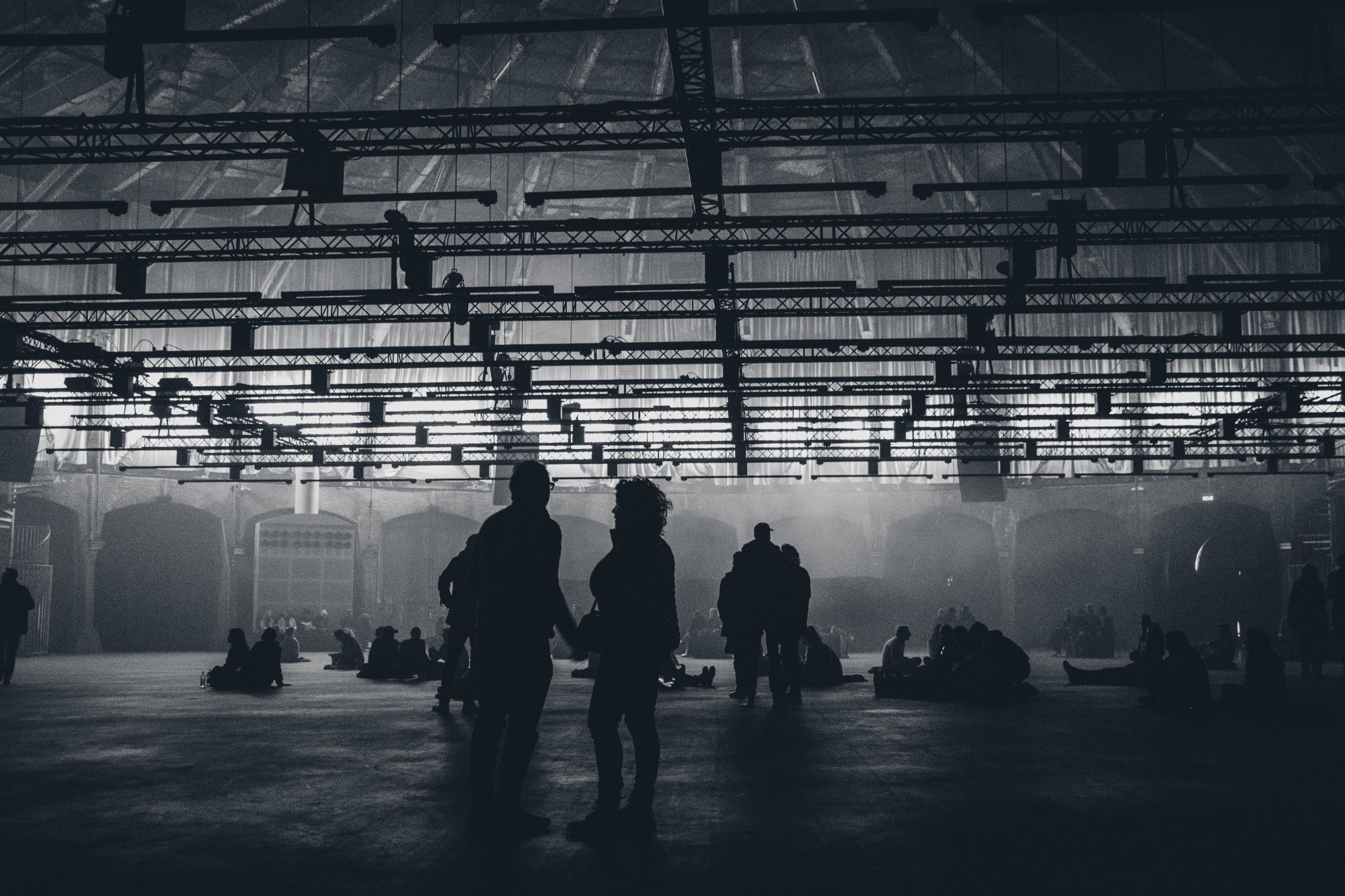 Silhouetten van mensen bewegen door een grote, industriële hal met een stalen dakconstructie, badend in diffuus licht en een mistige sfeer.