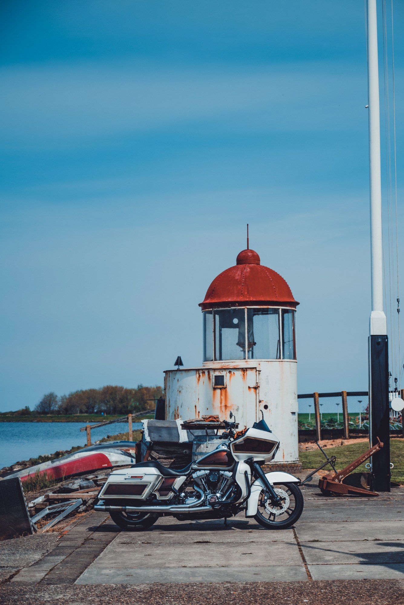 Een witte touringmotor staat geparkeerd op een kade bij een oude, verweerde vuurtoren met rode koepel, met daarachter water onder een strakblauwe lucht.