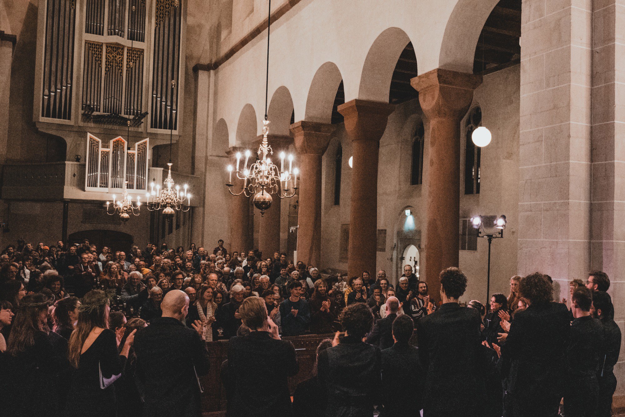 Publiek en koorleden applaudisseren in een sfeervol verlichte kerk met romaanse bogen, een groot orgel en hangende kroonluchters.