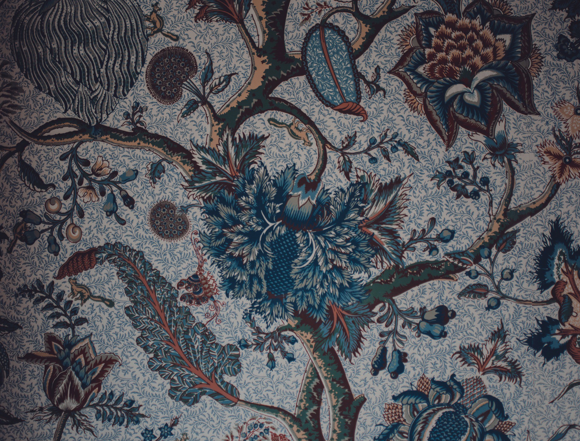 Rijk gedecoreerd textiel met een bloementak vol donkerblauwe en bruinrode bloemen, bladeren en knoppen tegen een fijn gebloemde lichte achtergrond.
