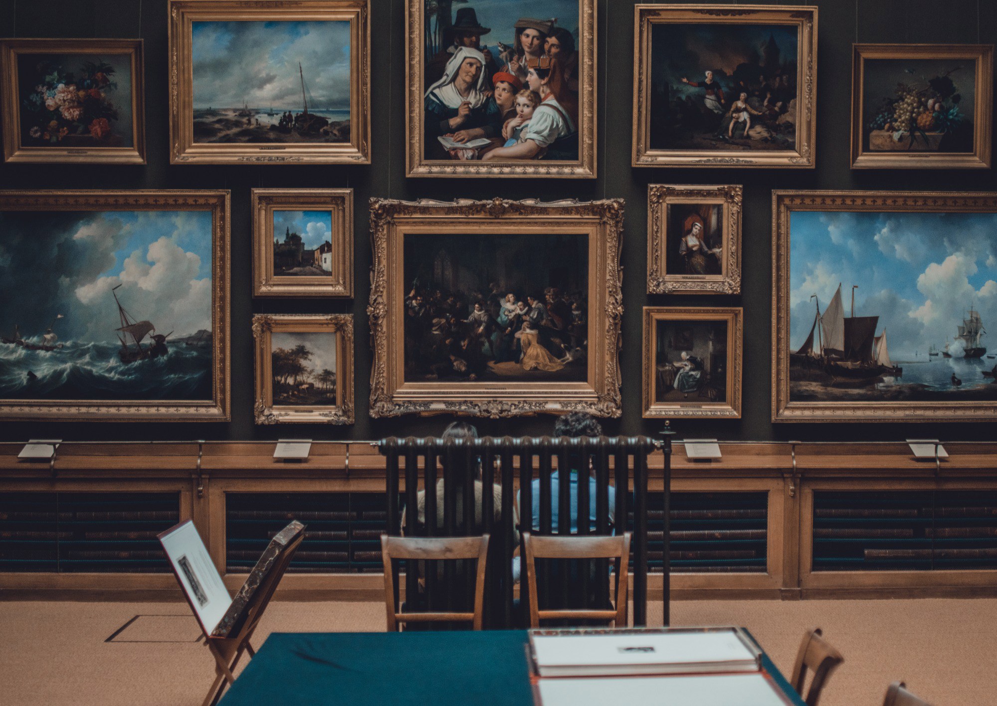Museumzaal met donkergroene wanden vol vergulde schilderijen in klassieke lijsten, met houten stoelen en een tafel met groen kleed op de voorgrond.