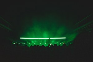 Een foto van achterin de zaal van Paradiso. We zien het publiek van achteren en de silhouetten van Weval. Ze worden van achter groen belicht door een streep op een LED-scherm.