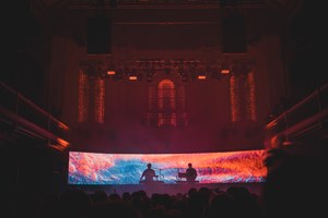 Een foto van achterin de zaal van Paradiso. We zien het publiek van achteren en de silhouetten van Weval. Ze worden van achter rood en blauw belicht door een visual op een LED-scherm.