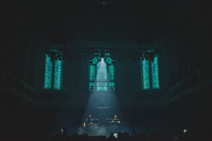 Een foto van achterin de zaal van Paradiso. Een spot richt van bovenin de zaal op het podium. We zien de kerkramen van Paradiso.