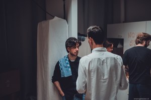 Twee jonge mannen staan in gesprek in een sfeervol verlichte studioruimte, terwijl op de achtergrond enkele andere personen bij een tafel bezig zijn.