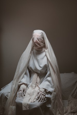 Een in witte doeken en een sluier gehulde figuur zit met de handen op de knieën op een bed, tegen een sobere bruine muur.