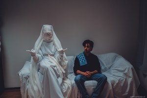 Twee personen zitten op een met witte lakens bedekte bank, waarvan één volledig in wit doek gehuld met bedekt gezicht en de ander in spijkerbroek toekijkt.