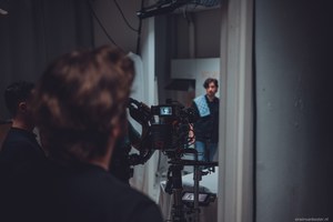 Op de voorgrond bedient een crewlid een filmcamera terwijl op de achtergrond een acteur met baard scherpgesteld in beeld staat.