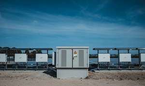 Een rij grijze technische kasten van een zonnepark staat opgesteld voor de zonnepanelen onder een strakblauwe lucht.