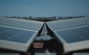 Twee rijen zonnepanelen lopen in een V-vorm naar elkaar toe onder een blauwe lucht, met in de verte windturbines en een bomenrij.
