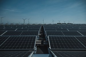 Rijen zonnepanelen strekken zich uit tot de horizon, waar windturbines onder een strakblauwe hemel oprijzen boen een groen heuvellandschap.