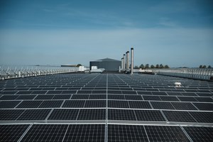 Een groot dak vol zonnepanelen strekt zich uit tot aan een grijze bedrijfshal met schoorstenen, onder een strakblauwe lucht.