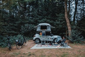 Op een beboste kampeerplek staat een auto met opgezette daktent, ernaast een fiets en een persoon op een campingstoel op een picknickkleed.