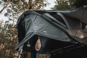 Een donkergrijze Thule Approach-daktent staat opgeklapt op een voertuig in het bos, met een hangende lamp en een petje aan haken en een ladder ernaast.