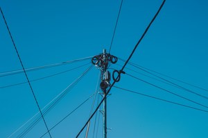 Een houten telefoonpaal met opgerolde kabels en isolatoren waaruit talloze draden uitwaaieren tegen een strakblauwe hemel.