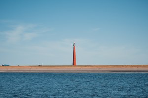 Een roodgekleurde vuurtoren staat op een smalle zandstrook tussen het water en de strakblauwe lucht, gezien vanaf de overkant van het water.