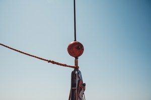 Een verweerde roestbruine haakblok met kraanhaak hangt aan een kabel tegen een strakblauwe lucht, met een gerafeld touw dat schuin het beeld in loopt.